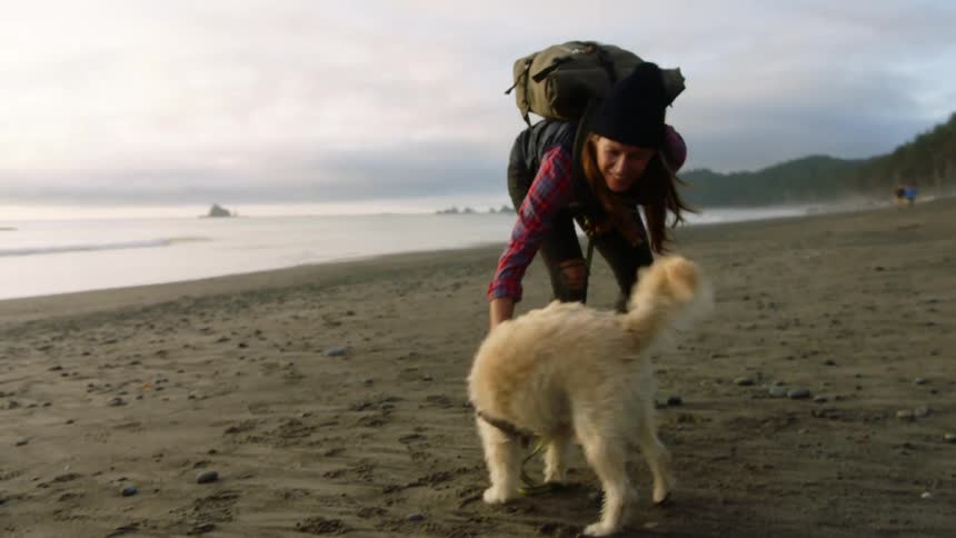 A woman in hiking gear plays with a dog on a beach as it fights with its leash