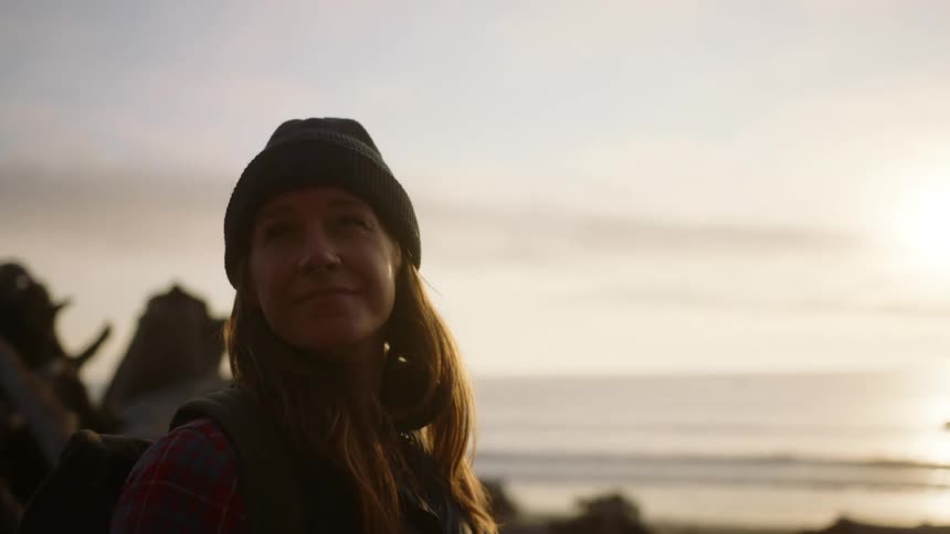 A woman in cold weather clothing looks around at her surroundings while standing on a beach littered with fallen trees during the day
