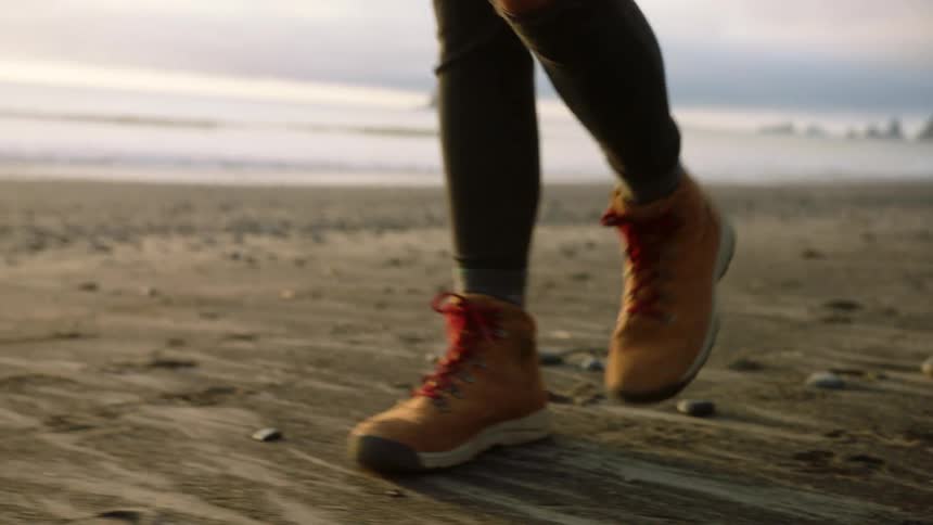 A woman with boots a backpack and a hat walking on the beach with the sky and clouds in the background