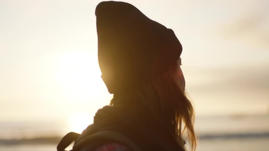 Woman with a black beanie and brown hair walks along the beach at sunset