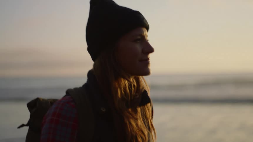 A woman in a winter cap smiling while walking on a beach at sunset