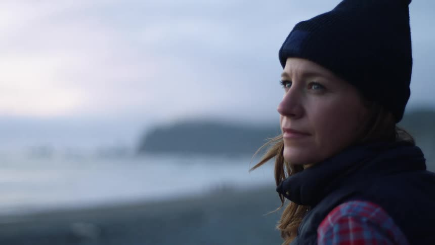 A middle aged woman in cold weather clothing stands on a beach while lost in thought on a cloudy day