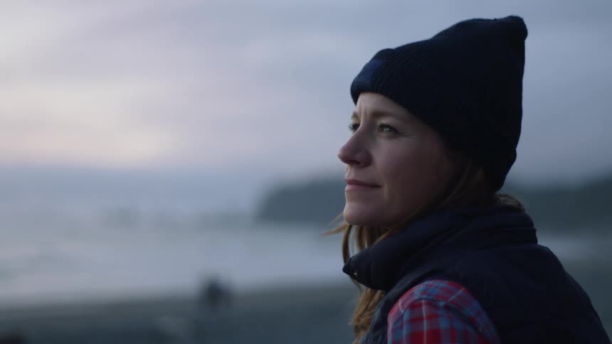 A woman in a winter hat standing on a beach and smiling at sunset