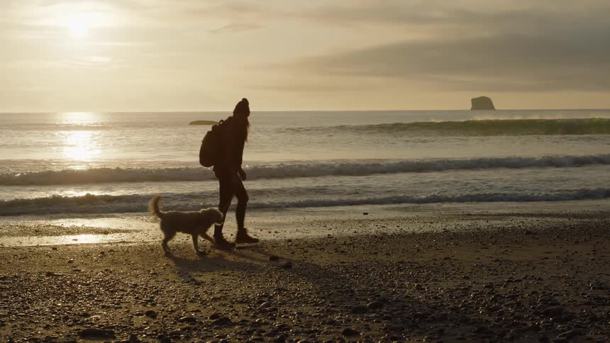 Woman with a beanie and a backpack walking a dog is silhouetted against the sunset on the pebble beach
