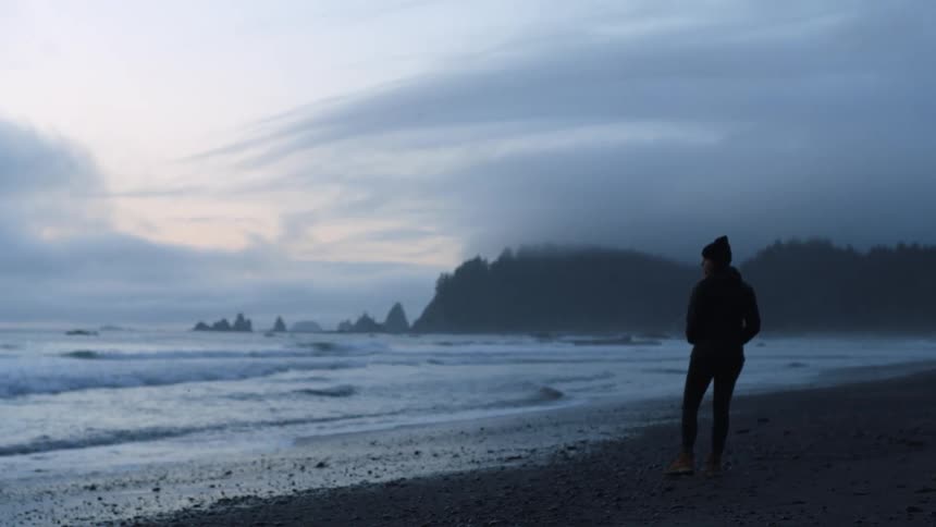 A person stands at the shoreline watching the water without a lot of sunlight wearing a hat and jacket