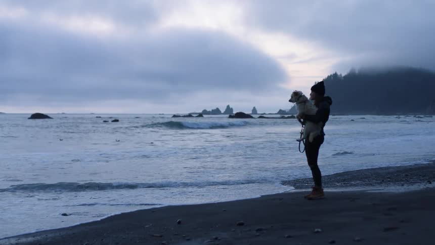 A woman holds her small white dog as she stands on the beach at the edge of the ocean