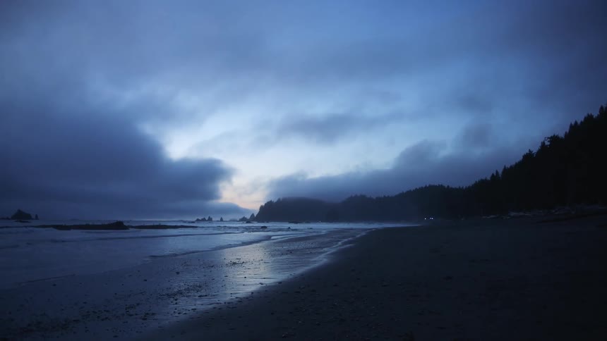 Moon shines through clouds onto beach bordered by forest as lit flickers in distance onshore