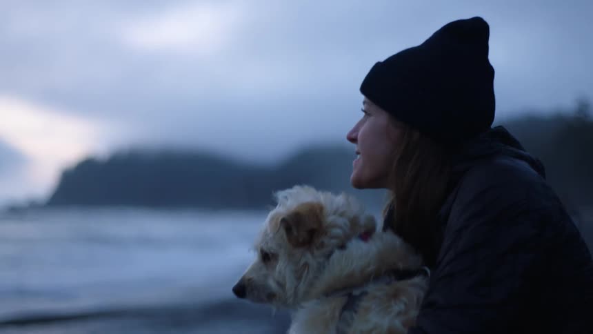 Woman with a black beanie and brown hair holds a white dog and holding out her finger towards the sea on the beach on a cloudy day