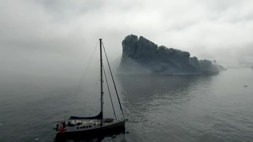Three people stand on a cruise ship with their coat hoods pulled up as the ship is stopped near an iceberg in the ocean