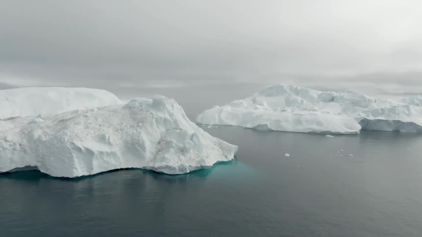 Several large ice bergs float atop the ocean water under a cloudy sky
