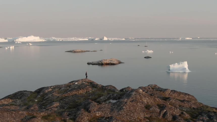 A person stands on the top of a mountain overlooking a body of water with ice flows