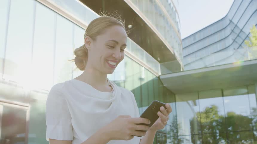 A woman stands outside an office building and smiles as she looks at her cell phone