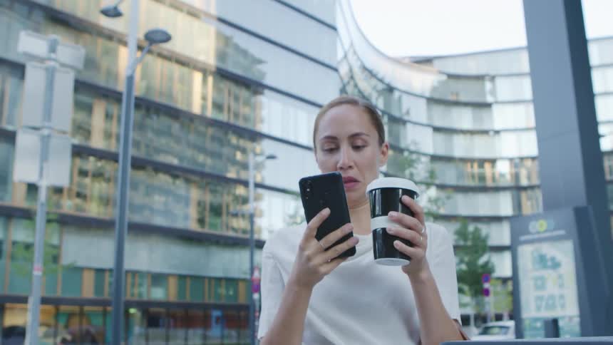 Woman stands near street talking on mobile phone then takes sip from portable coffee mug