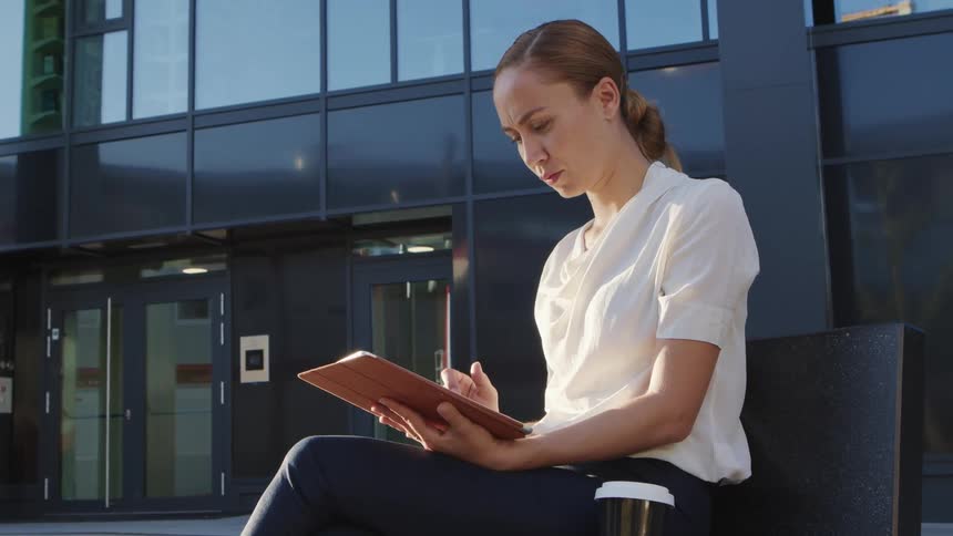A woman is sitting outside of a building while focused deeply on her tablet