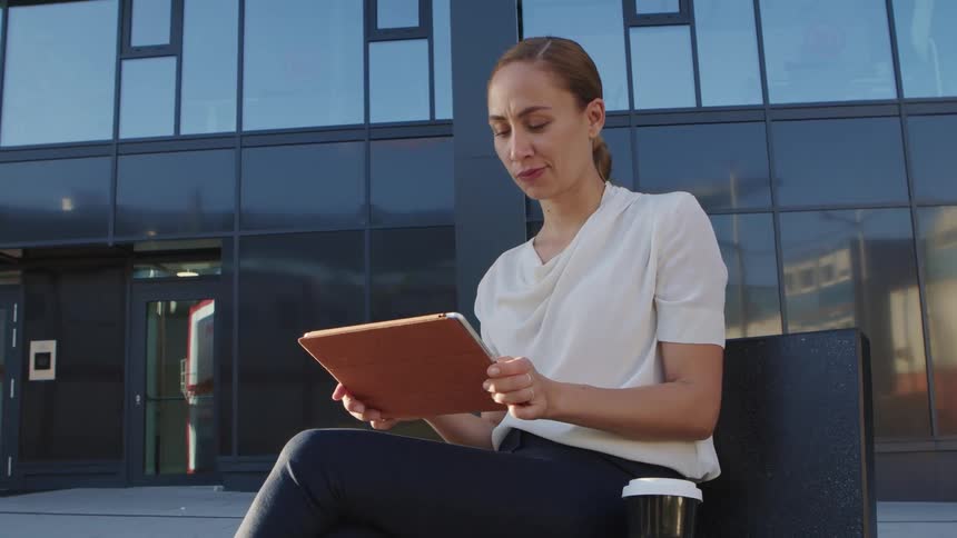 A woman sitting in front of a skyscraper looks up from her tablet and thinks for a moment before refocusing on her device