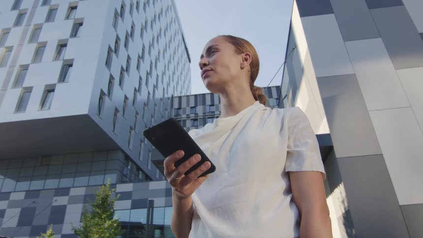 A woman stands outside of large city buildings holding a cell phone