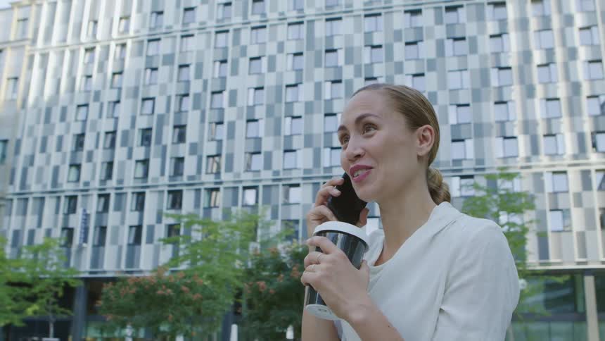 A young woman stands outside an office building in the city and talks on a cell phone and drinks a coffee