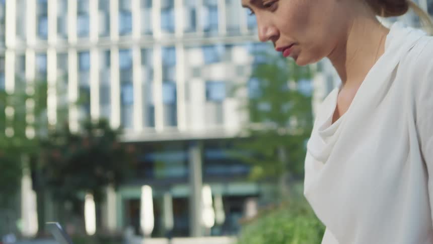 A woman uses a laptop while sitting outside of a skyscraper on a sunny day