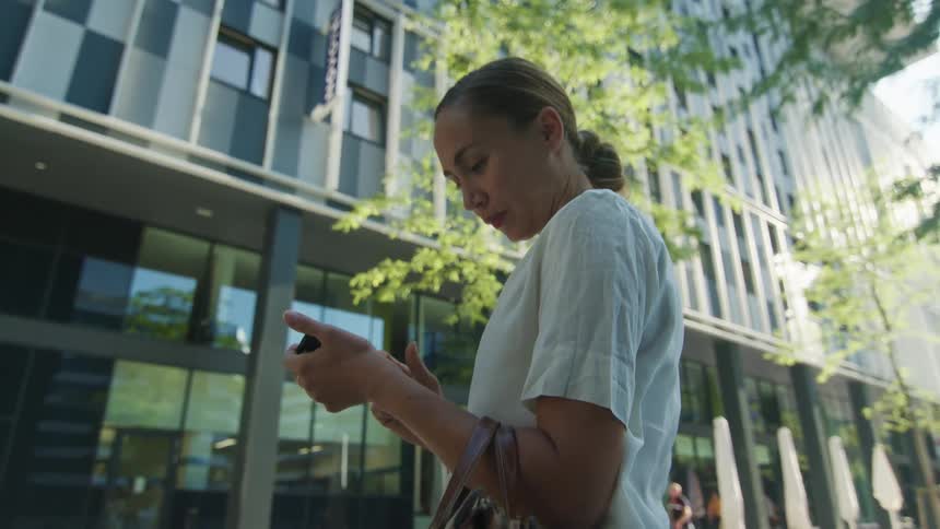 Woman with a white blouse holds a handbag and uses a smartphone in front of an office building