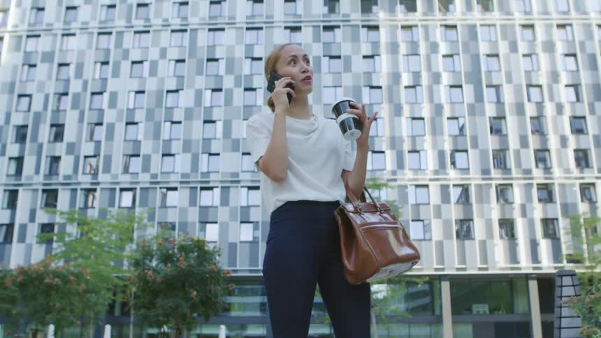 A woman talks on her smartphone while standing outdoors holding her purse and a cup of coffee in a metropolis during the day
