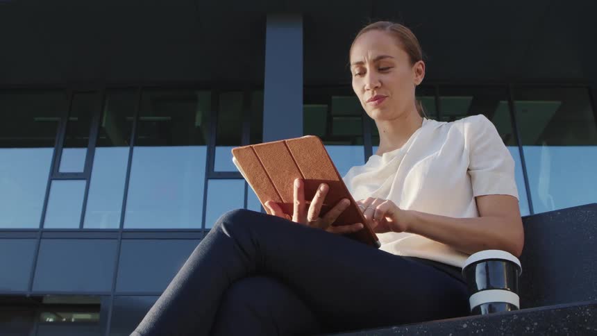 Businesswoman with a white blouse and black pants uses a tablet in front of an office building