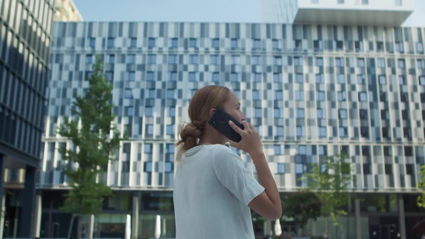 A woman stands in front of an office building talking on her cell phone on a sunny clear day
