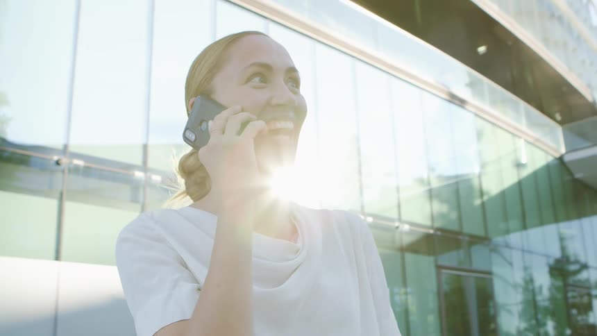 Woman stands near building reflecting sun talking on mobile phone and smiling