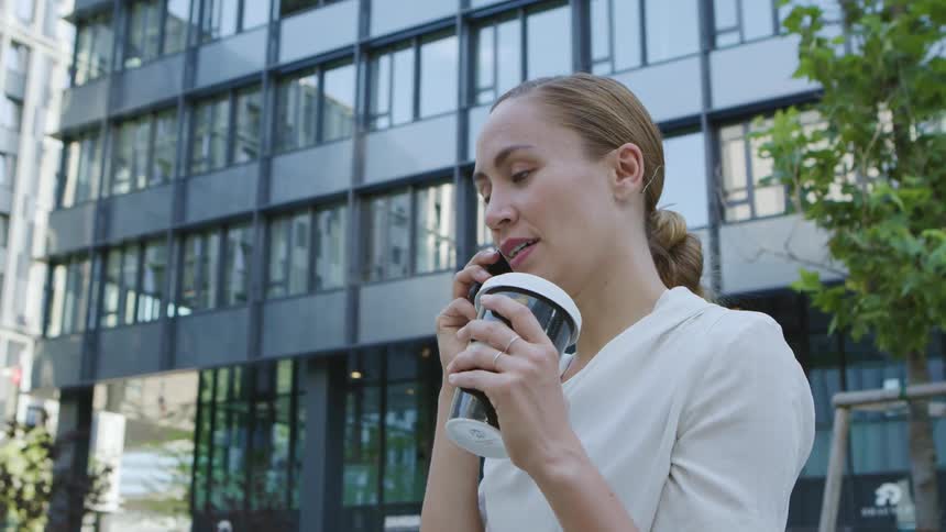 Woman with a white blouse holds a plastic coffee cup and talks on a smartphone in front of the office buildings