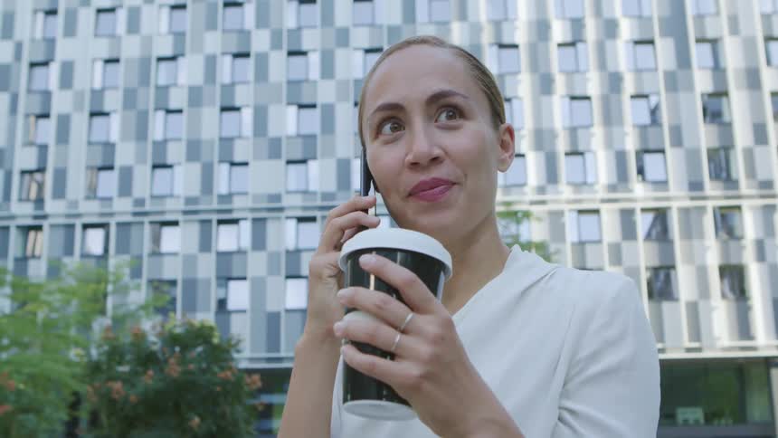 A woman sits outside an office building while talking on a cell phone and smiling while holding a coffee cup