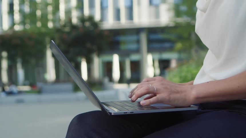 A person sitting and typing on a laptop with trees and a building in the background