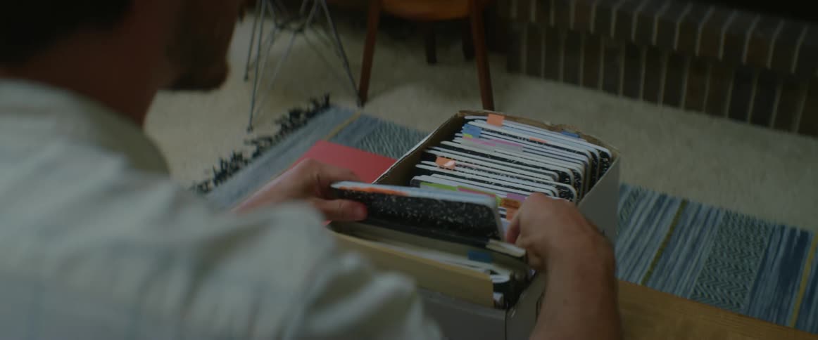 A man sits on a couch and looks through a box of notebooks pulling one out and rifling the pages in a family room