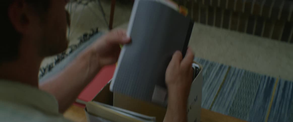 A man pulls a notebook out of a box filled with notebooks flips through the pages and places it back in the box on a table in a living room