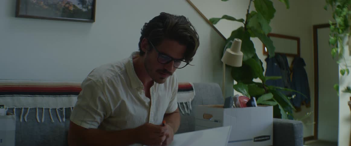 Man with glasses a white shirt sits in a couch and writes on a white board in the living room at home