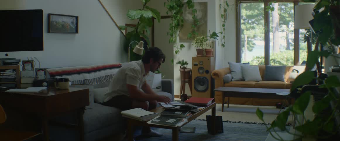 Man sitting on couch in living drinking from a mug then cleaning a spill on his shirt