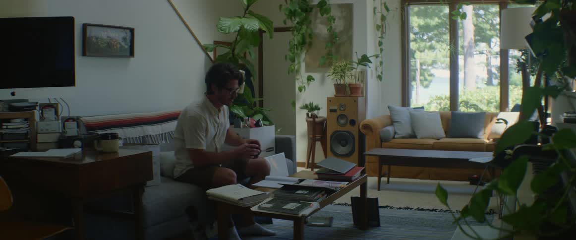 A man walks in to his living room and sets his coffee on a table and looks at papers