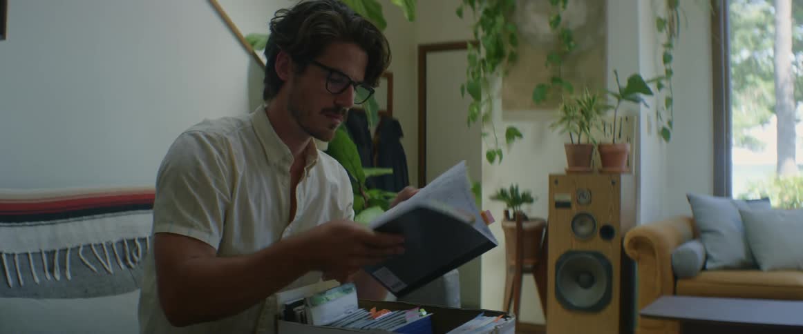 A man sits on his couch and looks through a box and pulls out a notebook and sets it aside