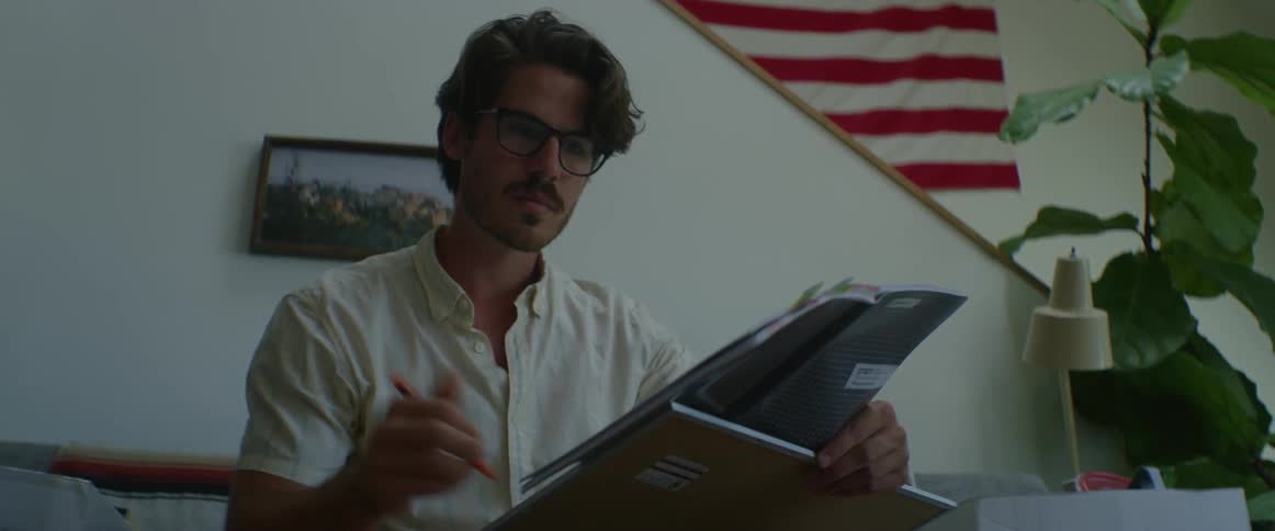 Man with glasses a white shirt sits in a couch and writes on a whiteboard in the living room at home