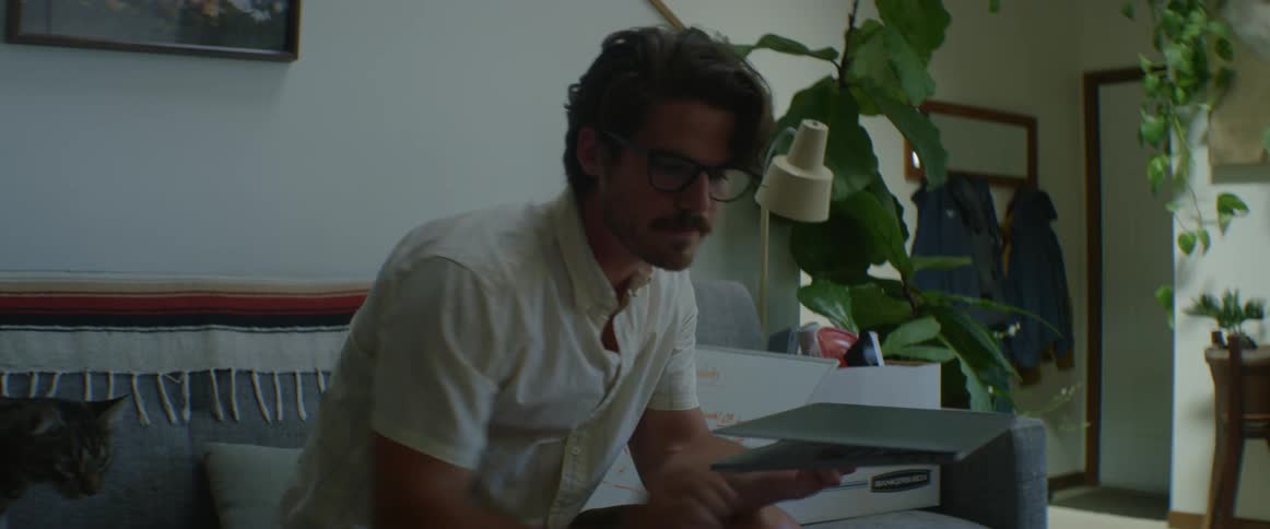 A young man at home picks up his laptop takes a drink of coffee and then opens his lap and sets it down and picks his cat up