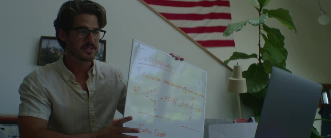 A man holds a whiteboard up to a laptop camera as he speaks while participating in a video conference