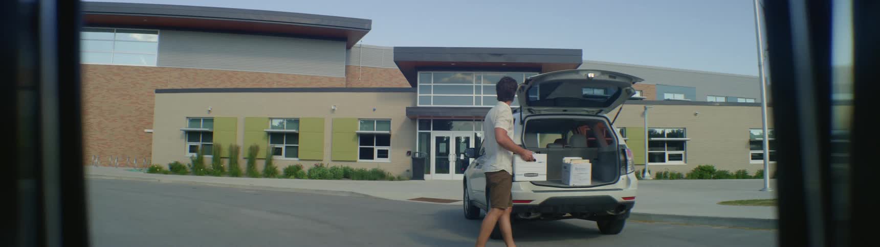 A man carries a box of files and loads it into his car parked in the empty driveway of a college building
