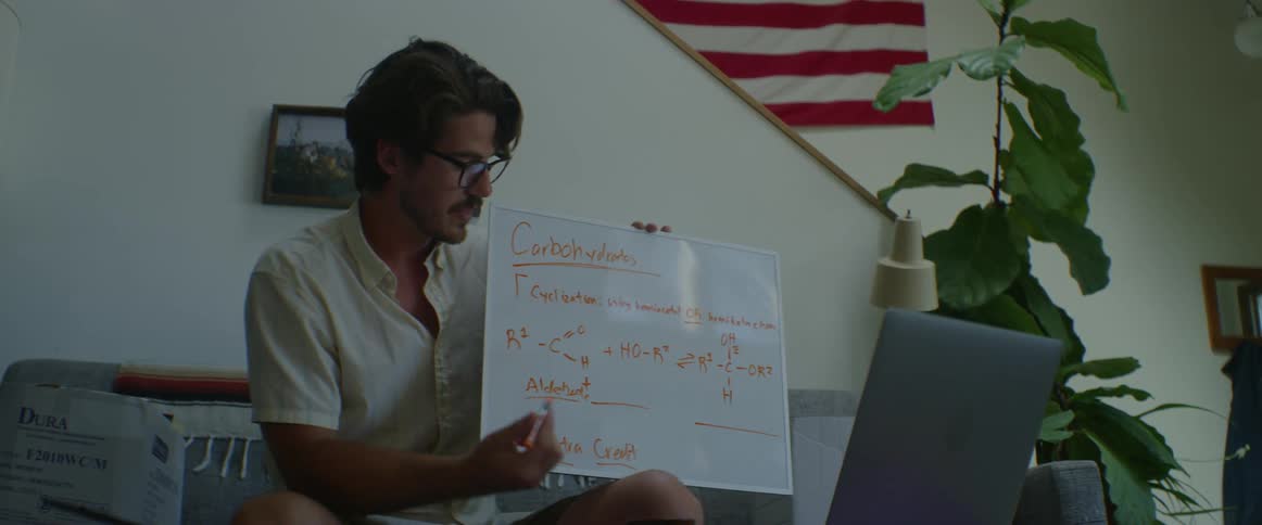 A man sits on a table holding a white board writing and explaining in a room