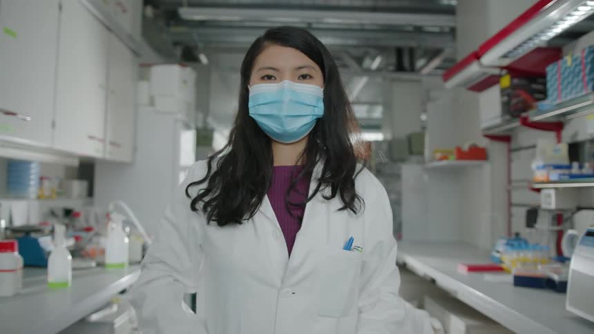 A woman wears a lab coat and face mask in a white lab setting brushing her hair forward over her shoulder