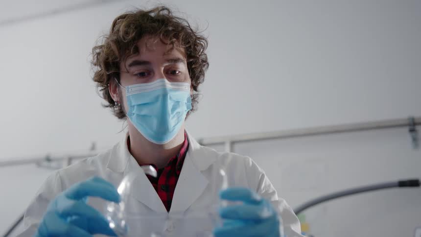 A man with gloves a mask and a lab coat taking off goggles