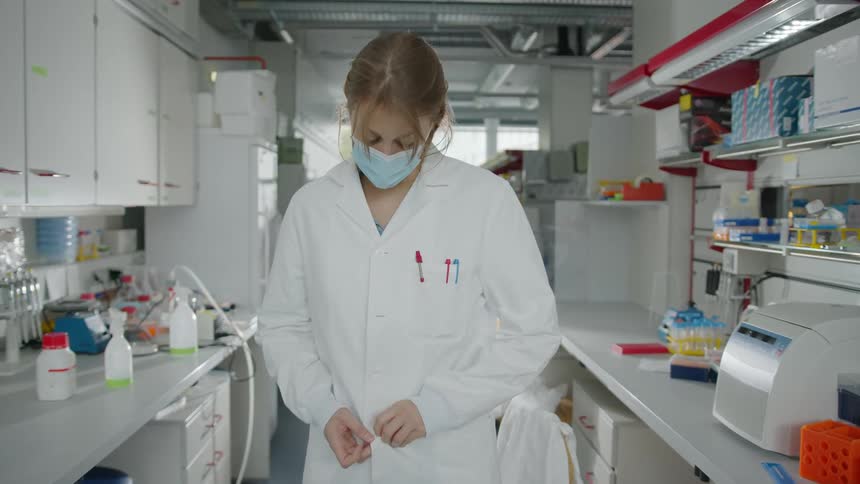 A blonde woman is buttoning her lab coat and placing her hands in the pockets while wearing a face mask in a lab setting