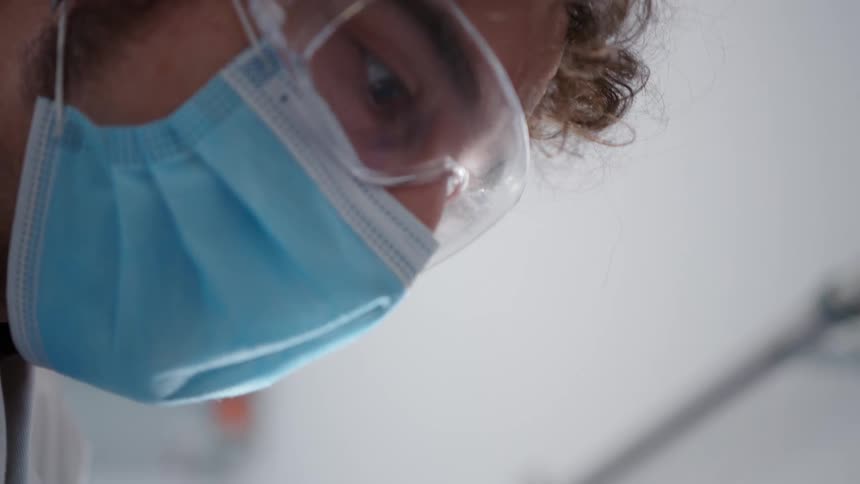 A man wearing a medical mask and safety glasses focuses on something as fog mists by his face