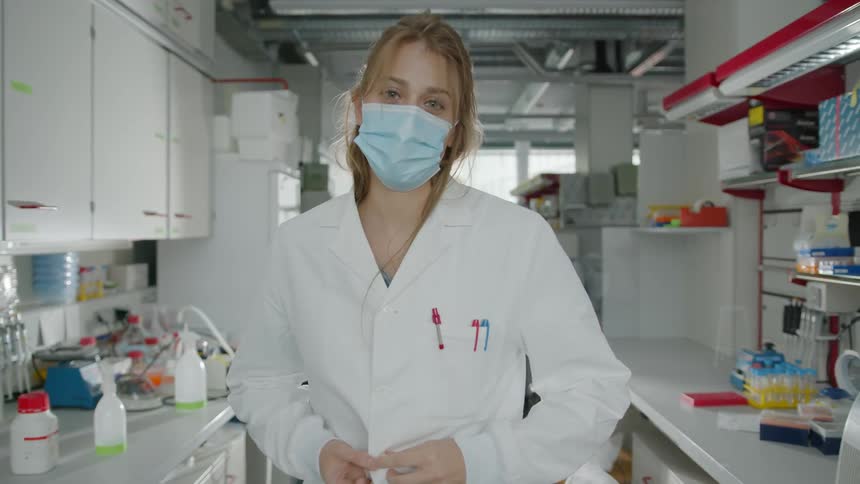 A woman with a mask and a lab coat standing and staring in a lab