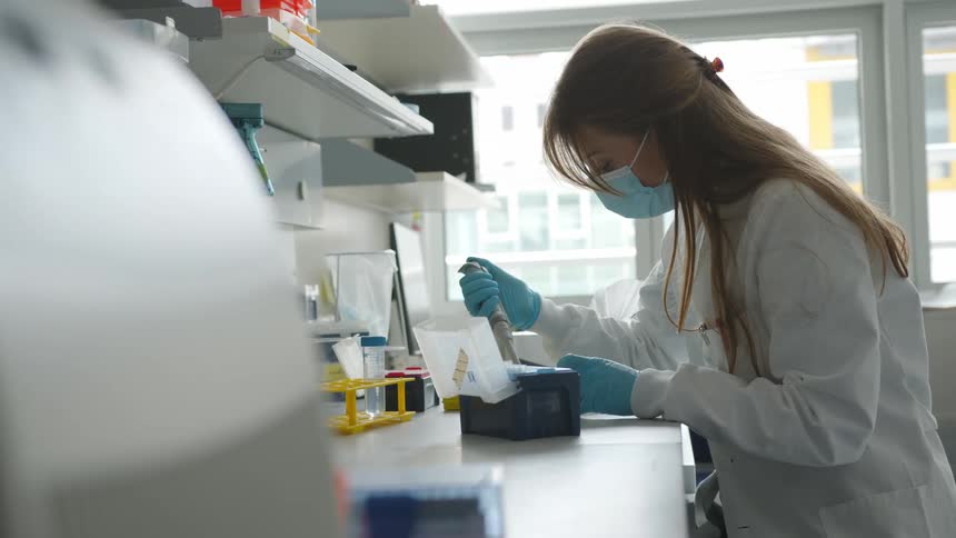 A woman wearing a lab coat face mask and gloves samples liquid in a lab setting while sitting on a stool in daytime