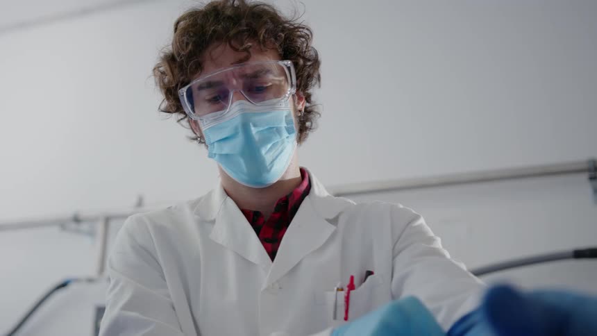 Man wearing a lab coat standing in lab putting on goggles and gloves