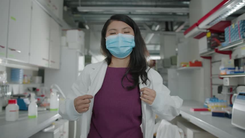 Woman wearing face mask dons white lab coat standing between counters holding laboratory supplies