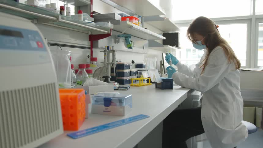 A woman in a lab coat face mask and gloves sits on a stool while working in a lab during daytime